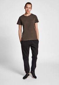 Olijfgroen katoenen T-shirt met een ronde hals, gecombineerd met zwarte taps toelopende broek. Model draagt zwarte schoenen en staat met handen in de zakken.