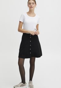 Jupe A-line en daim noir avec fermeture à boutons à l'avant et deux poches latérales, associée à un T-shirt blanc ajusté et des collants noirs.