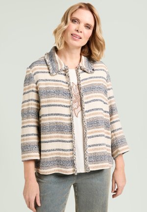 Blondynka ubrana w beżową, niebieską i brązową, prążkowaną, teksturowaną kurtkę na białym topie i niebieskich dżinsach, stojąca na jasnym, jednolitym tle.