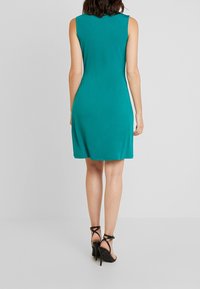 Ärmlös teal klänning med en mjuk textur, som faller ovanför knäna och har en figursydd design. Bärs med svarta sandaletter med hög klack.