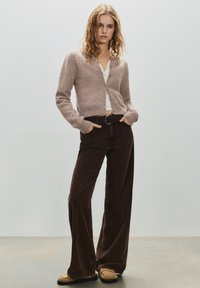 Femeie cu păr blond ondulat, purtând un cardigan maro deschis cu nasturi, un top alb, pantaloni largi din catifea cu dungi, de culoare maro închis, și pantofi loafer bej.