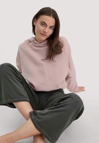 Vrouw met lang bruin haar die in kleermakerszit zit, gekleed in een lichtroze hoodie en donkergroene wijde broeken tegen een eenvoudige achtergrond.