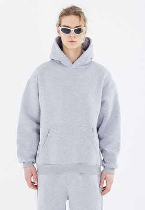 Grauer Hoodie mit einer Fronttasche, gerippten Bündchen und einem entspannten Schnitt. Getragen mit weißen Cat-Eye-Sonnenbrillen; weiche Textur, minimales Branding.