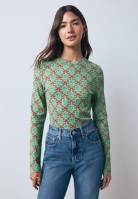 Haut à manches longues en coton vert texturé, arborant un motif floral rouge et bleu répété, associé à un jean en denim bleu taille haute.