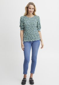 Blusenshirt mit floralem Muster in Blau- und Grüntönen, mit Puffärmeln; kombiniert mit enganliegenden blauen Jeans und schwarzen Loafers.