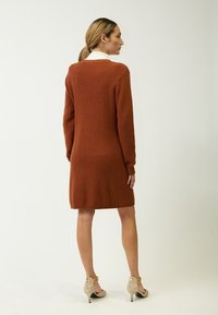 Vestido suéter de longitud hasta la rodilla en color óxido con textura de canalé, mangas largas y un sutil escote en V. Combinado con tacones beige con acentos met�álicos.