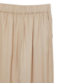 Un pantalon beige à taille élastique présente une texture lisse et brillante avec des plis et des poches latérales, taillé pour une coupe ample.