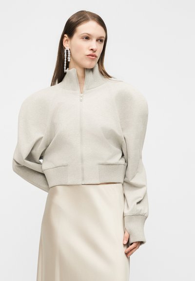 Femme portant un pull beige clair à fermeture éclair avec des manches bouffantes et une jupe en satin beige, accessoirisée avec de longues boucles d'oreilles pendantes.