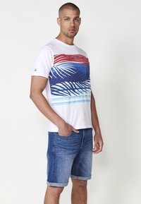 Balta īsās piedurkņu T-krekls ar palmu lapu grafiku sarkanā, zilā un tirkīza krāsā, savienots ar sarullētām zilajām denim šortiem.