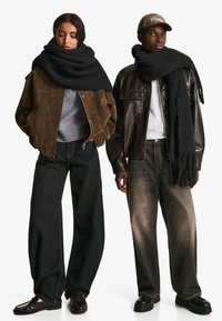 Jachetă bomber din suede maro, pulover tricotat gri, blugi negri cu picioare largi și eșarfă neagră pe o femeie; jachetă din piele neagră și blugi închiși la culoare pe un bărbat.