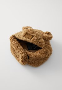 Chapéu de urso de pelúcia na cor parda, feito de um tecido macio e texturizado, com orelhas arredondadas e um rótulo no interior. O interior é escuro e liso.