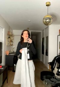 Kvinde med mørkt hår holder lysegrå bukser med mærker i en moderne stue med guldfarvede hylder, en globus-pendel og en gang i baggrunden.
