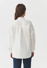 Bílá dlouhá košile s límečkem, zadním skladem a knoflíkovými manžetami, vyrobená z lehké texturované látky. Stylizováno s tmavými džíny.