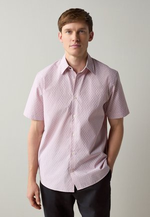Homme debout devant un fond uni portant une chemise rose clair à manches courtes avec un motif géométrique subtil et un pantalon noir.