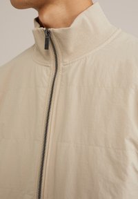 Giacca zip-up beige chiaro con colletto alto, tessuto strutturato e zip nera. Presenta cuciture orizzontali e una finitura liscia.