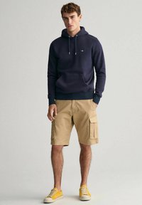 Marineblå hættetrøje med frontlomme, parret med khaki cargo shorts med sidelommer. Gule snørebåndssneakers fuldender outfittet.