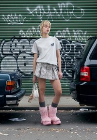 Hall halli T-särk logoga, mustriline miniseelik, roosad karvased saapad, hallid sokid, hoides väikest kotti ja pehmet mänguasja, graffitikunsti tausta ees.