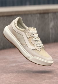 Beige sneakers med teksturert stoff og glatte lærdetaljer, med hvit såle og tonede lisser, viste flytende mot en nøytral bakgrunn.