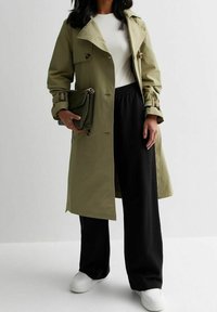 Olivově zelený trenčkot s knoflíkovými detaily, nošený přes bílý žebrovaný top a černé široké kalhoty, doplněný zelenou kabelkou a bílými teniskami.