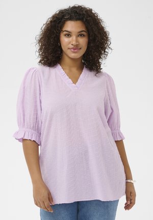 Kobieta o kręconych włosach, ubrana w teksturowaną liliową bluzkę z dekoltem w serek i bufkami oraz niebieskie dżinsy, stojąca na jednolitym białym tle.