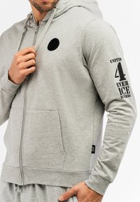 Sivá mikina na zips vyrobená z mäkkej látky s prednou vreckom, čierna nášivka s logom na hrudi a potlačený text pozdĺž rukávu.