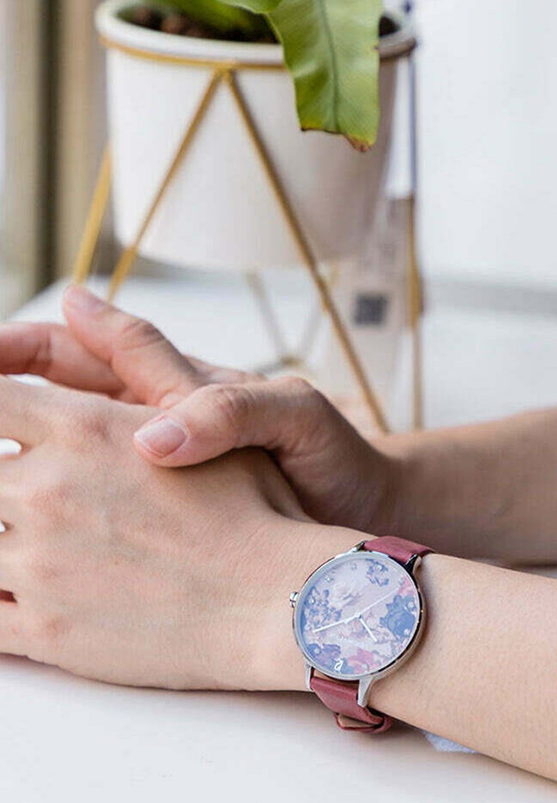 Montre ronde avec un boîtier métallique, un cadran à motif floral et un bracelet en cuir bordeaux. Aiguilles reposant sur une surface, avec une plante en arrière-plan.