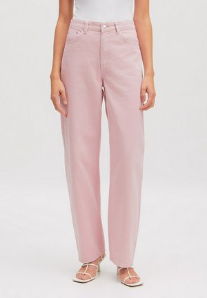 Frau trägt hochtaillierte, weit geschnittene hellrosa Jeans kombiniert mit weißen, riemchenbesetzten Sandalen mit Absatz vor einfarbigem Hintergrund.