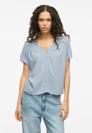 Lichtblauwe blouse met korte mouwen en een V-hals, voorzien van decoratieve stiksels langs de naden, gecombineerd met lichtblauwe denim jeans.