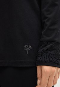 Zwarte longsleeve top met getextureerde kanten accenten op de mouwen en een subtiele reliëflogo op de zoom, gemaakt van een zachte, rekbare stof.