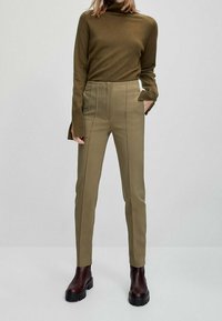 Olijfgroene coltrui gecombineerd met hooggewaisted beige broeken. De broeken hebben een flat front met gestikte details; het model draagt donkerbruine laarzen.