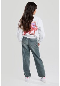 Felpa grigia con testo grafico rosso, abbinata a jeans in denim sbiaditi e dal taglio morbido. Il modello indossa sneaker bianche con dettagli rossi.