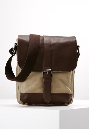 Bolso pequeño de lona beige con solapa de cuero marrón oscuro, correa con hebilla y correa de hombro de tela ajustable sobre superficie blanca.