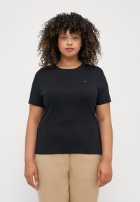 Svart kortärmad t-shirt med rund halsringning och en liten logopatch på bröstet, ihop med beige byxor. Slät tygtextur.