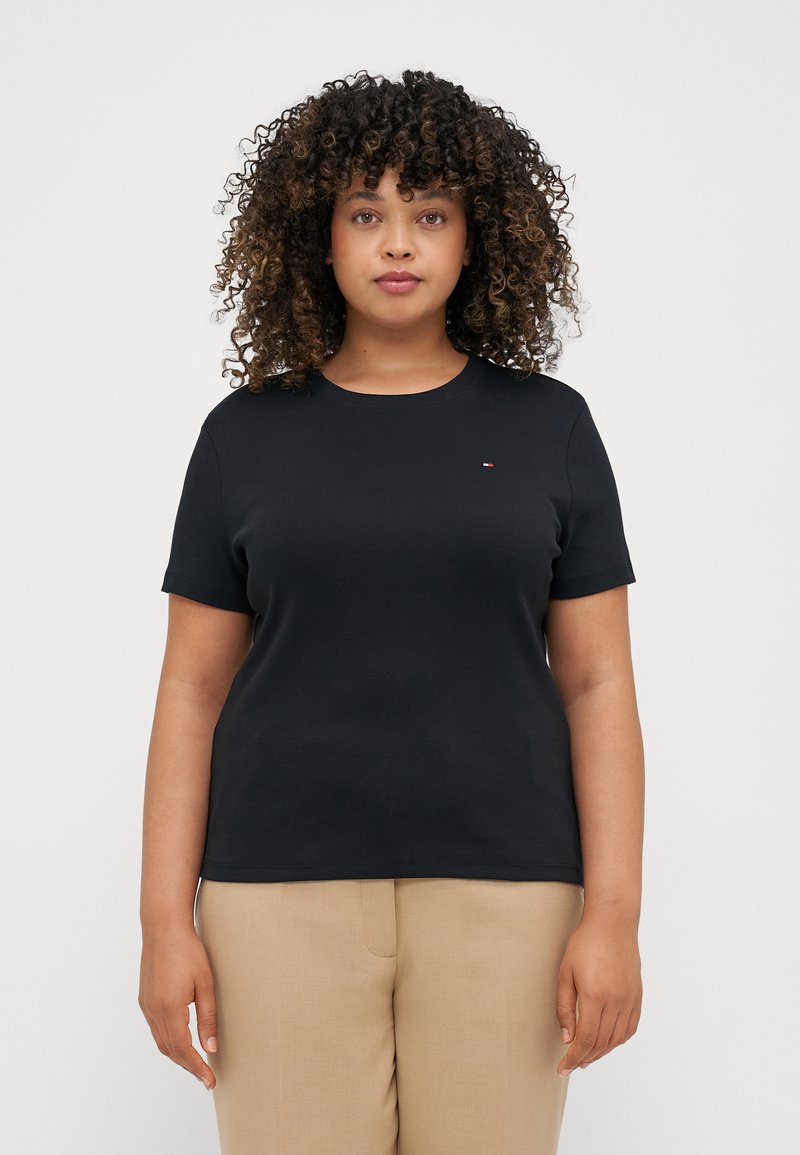 Svart kortärmad t-shirt med rund halsringning och en liten logopatch på bröstet, ihop med beige byxor. Slät tygtextur.