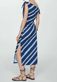 Robe rayée bleu marine avec des accents bleu clair, design asymétrique, bretelles à nouer et une fente latérale. Portée avec des sandales à lanières noires.