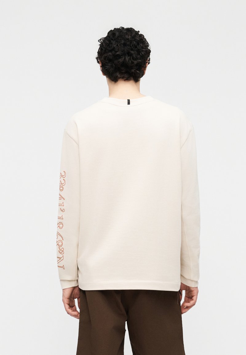 Person with curly dark hair wearing a cream long-sleeve shirt with brown pants, back facing camera, standing against a plain light background.