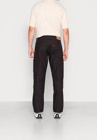 Pantalones cargo negros con múltiples bolsillos, un diseño de pierna recta y una etiqueta de marca visible en la cintura; combinados con zapatillas blancas.