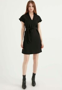 Schwarzes Kleid mit V-Ausschnitt, kurzen Ärmeln und gebundenem Taillenausschnitt. Aus glattem Stoff gefertigt, mit geradem Saum und kombiniert mit schwarzen Stiefeletten.