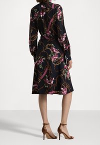 Svart klänning med blommig kedjemönster, långärmad och med markerad midja. Bärs med bruna öppna högklackade skor. Slät tygstruktur.