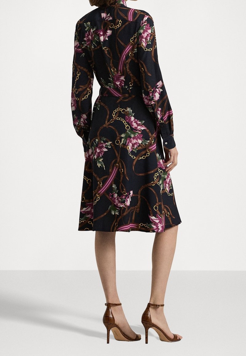 Svart klänning med blommig kedjemönster, långärmad och med markerad midja. Bärs med bruna öppna högklackade skor. Slät tygstruktur.