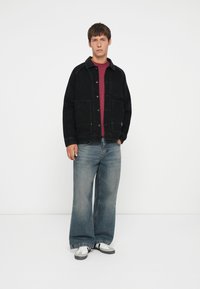 Black denim jacket with front pockets, paired with a maroon t-shirt and wide-leg blue jeans. White and gray sneakers complete the look.