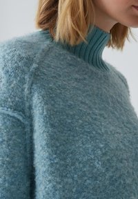 Gros plan sur une personne portant un pull en maille bleu clair texturé avec un col montant côtelé et des cheveux blonds aux épaules.
