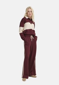 Tuta bicolore bordeaux e crema con polo a maniche lunghe e pantaloni con coulisse. Presenta strisce laterali e una vestibilità comoda. Texture morbida.