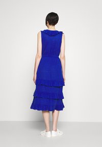 Blå, ärmlös klänning med rynkad hals och flerlagers volang nertill, tillverkad av strukturerad tyg. Vita slip-on skor fulländar looken.