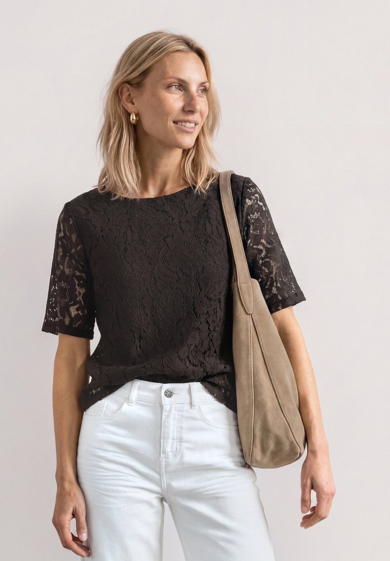 Femme blonde portant un top noir en dentelle à manches courtes, pantalon blanc, boucles d'oreilles en or et portant un sac à épaule beige, regardant vers la droite.