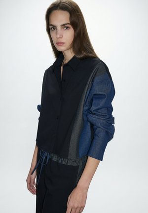 Młoda osoba z długimi brązowymi włosami, ubrana w czarną i niebieską koszulę na guziki z podwiniętymi rękawami, stojąca na jasnym, jednolitym tle.