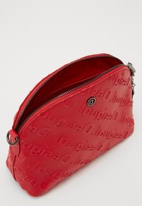 Sac bandoulière rouge avec logo de la marque en relief, texture lisse et fermeture éclair argentée. Dispose d'une boucle latérale pour attache.
