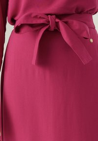 Abito fucsia con vita regolabile e texture di tessuto liscia. Include un piccolo bottone dorato come dettaglio aggiuntivo.