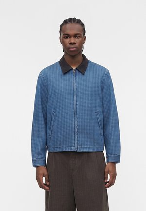 Jeune homme aux cheveux tressés portant une veste zippée à carreaux bleus avec un col noir et un pantalon ample marron foncé sur fond blanc.