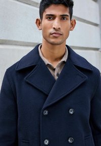 Abrigo de lana mezcla en color navy con diseño de doble botonadura, grandes solapas y cuatro botones negros. Lleva una camisa de color beige con cuello debajo.